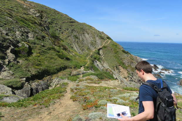 A person with a backpack holds a map while standing on a rocky coastal path. The path winds up a hill beside the ocean under a clear blue sky.
