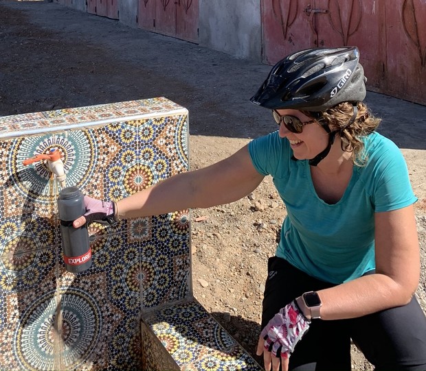 A person in a bike helmet fills a bottle labeled "EXPLORE" from a mosaic-tiled fountain, smiling in a sunny outdoor area.