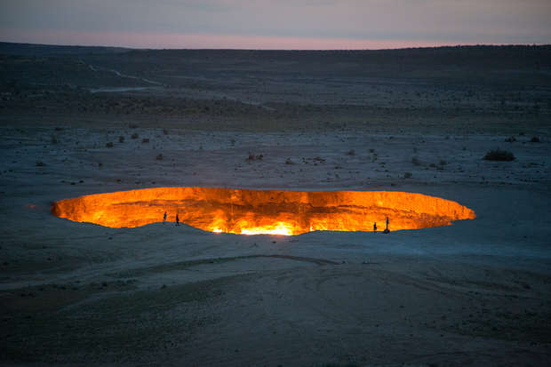 A large, fiery crater emits bright orange light, surrounded by people standing on the rim, in a barren desert landscape under a dim sky.
