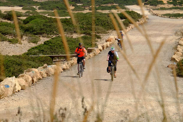 Cyclists ride along a gravel path lined with rocks, surrounded by green shrubs, under an open sky. A couple leads while others follow, creating a scenic outdoor biking scene.