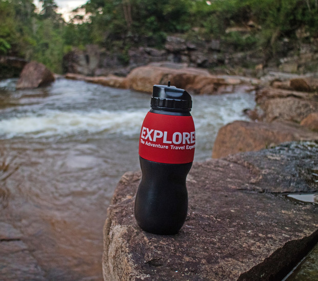 A black water bottle with a red label reading "EXPLORE - The Adventure Travel Experts" sits on a rock by a flowing river surrounded by greenery.