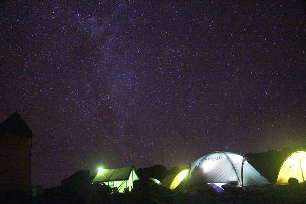 Tents are illuminated against a starry night sky, set in a clear outdoor camping environment. The Milky Way is visible overhead, adding to the serene atmosphere.