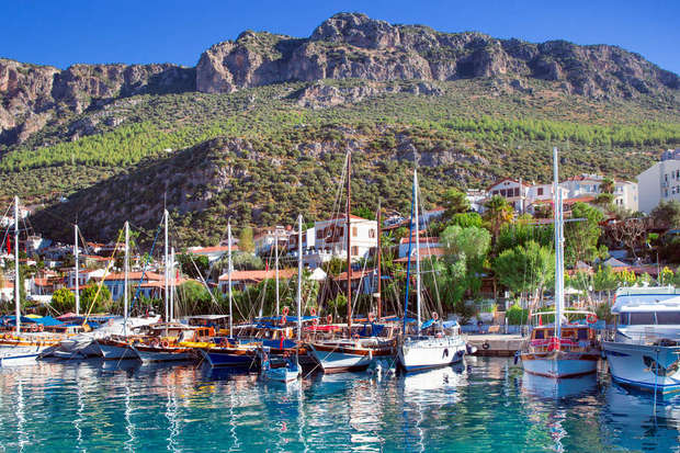 Boats are docked in a marina set against a backdrop of green hills and rocky mountains. The scene includes houses with red roofs and lush greenery under a clear blue sky.