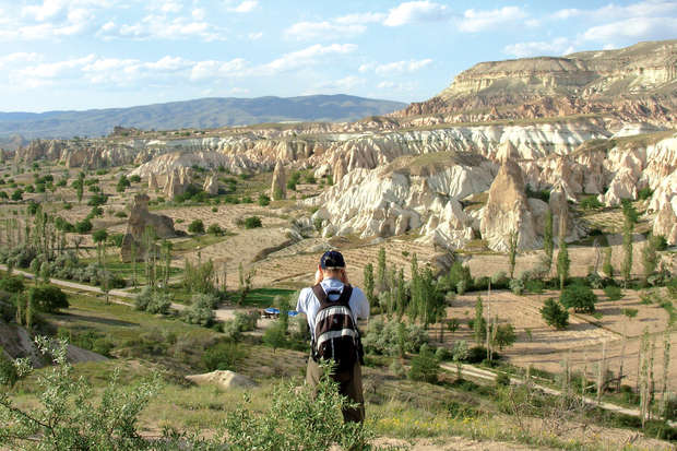 A person wearing a backpack stands on a grassy hill, photographing a vast landscape of rocky formations and green shrubs under a blue sky with scattered clouds.