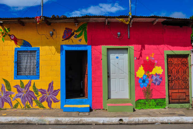 Colorful house with yellow and pink walls, decorated with floral and bird murals; blue and green door frames. Situated along a street with a clear blue sky above.
