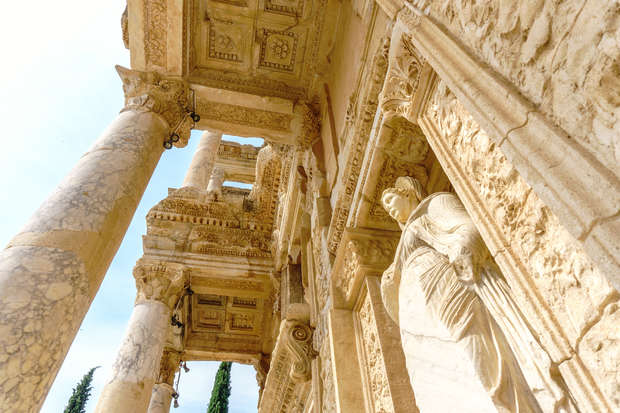 Ancient stone columns support an ornately carved facade featuring a classical statue, all under a bright sky. The structure's intricate details and textures evoke a historical architectural style.