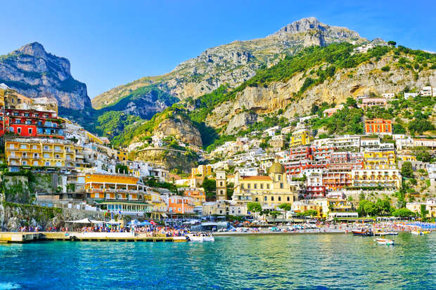 Colorful hillside buildings stacked against a steep mountain, overlooking the clear blue sea. People gather near the waterfront, with boats scattered on the water, completing the vibrant coastal scene.