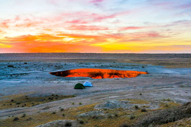 Two tents sit near a glowing, fiery pit reflecting the vibrant hues of a sunset in a barren, flat landscape, creating a dramatic and colorful scene.
