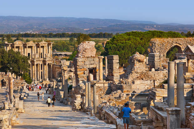 Ancient ruins line a wide stone street where tourists walk, surrounded by columns and partially standing structures. In the background, hills and greenery are visible under a clear sky.