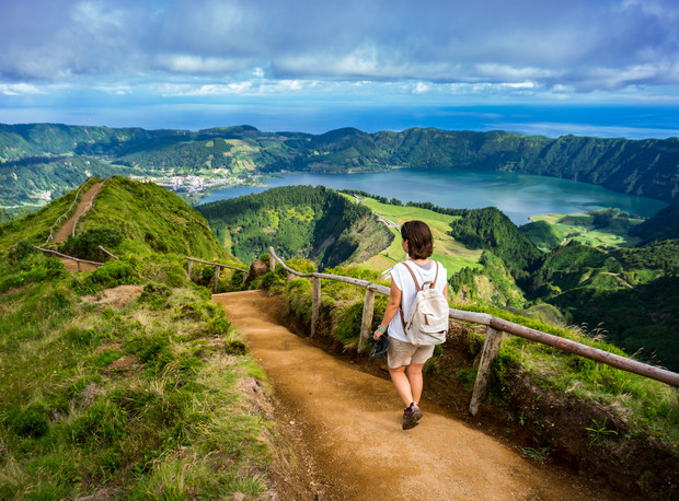 A hiker walks a winding dirt path surrounded by lush hills, overlooking a scenic lake and distant village under a partly cloudy sky.
