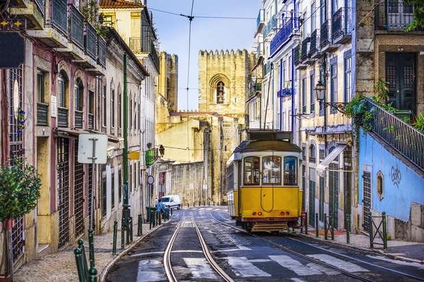 A yellow tram moves along tracks down a narrow, cobblestone street lined with colorful buildings in a historic European city, with an ancient stone cathedral visible in the background.