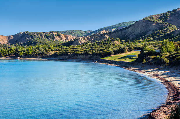 A serene bay features calm blue waters gently lapping against a curved sandy shoreline, bordered by lush greenery, with rugged, tree-covered hills rising in the background under a clear sky.