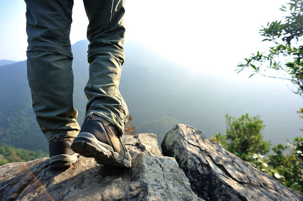 Hiking boots stand firmly on a rocky edge, overlooking a vast, misty mountainous landscape. Nearby, sparse greenery breaks the rocky terrain, suggesting a scenic viewpoint in a natural setting.
