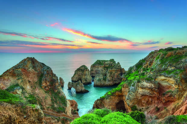 Rocky cliffs frame two sea arches, with lush greenery atop, while vibrant ocean waves gently crash below at sunset, blending pink, blue, and orange hues across the sky and water.