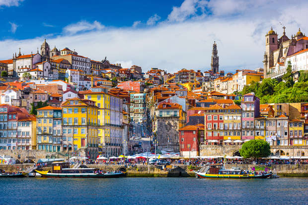 Colorful hillside buildings stand overlooking a busy waterfront with boats and people. The scene is set under a blue sky with scattered clouds, showcasing a vibrant cityscape.