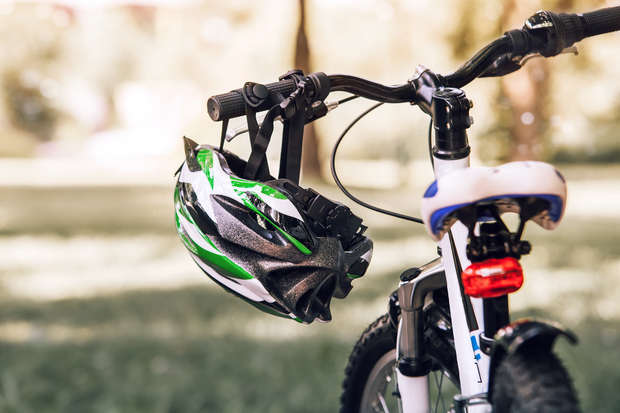 A bicycle stands in a grassy park, with a black and green helmet hanging from the handlebars. Sunlight filters through the surrounding trees, creating a dappled effect.
