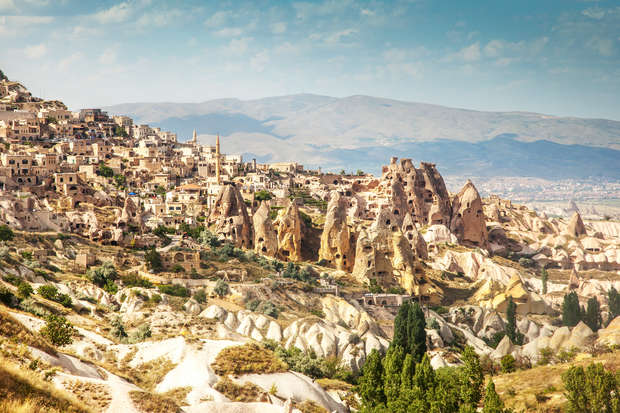 Rock formations rise amidst an ancient town with stone buildings, set against arid hills and a clear sky. Vegetation sparsely dots the foreground, enhancing the rugged landscape.