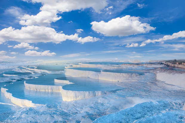 Terraced pools filled with blue water cascade down white travertine formations under a bright blue sky with scattered clouds, creating a dramatic natural landscape.