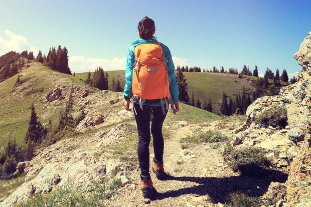 A person with an orange backpack hikes along a rocky trail surrounded by green hills and scattered pine trees under a clear blue sky.