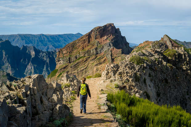 A person wearing a bright yellow backpack walks along a rocky mountain path, flanked by jagged cliffs, under a partly cloudy sky.