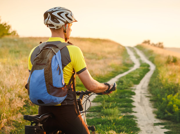A cyclist wearing a helmet and backpack rides a bike along a dirt path through grassy fields during sunset, creating a serene outdoor setting.