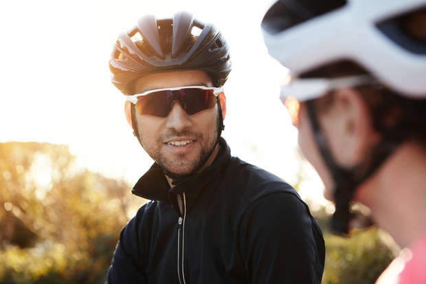 A person wearing a black helmet and sunglasses smiles while talking with another person in a similar outfit, outdoors with sunlight and foliage in the background.