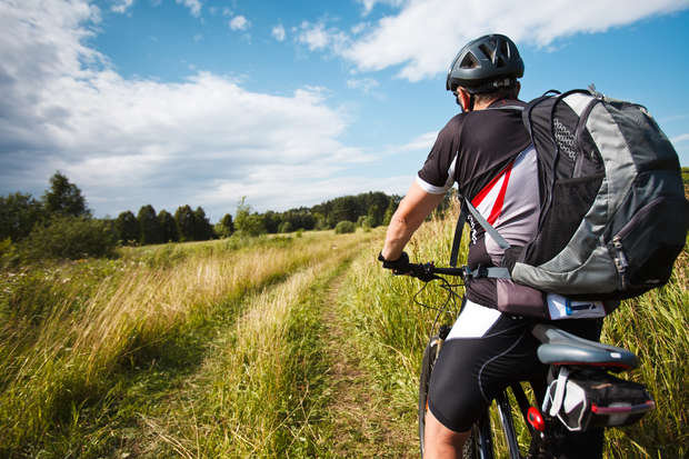 A cyclist rides along a grassy trail, wearing a helmet and backpack. The scene features a bright, open landscape with trees and a partially cloudy sky in the background.