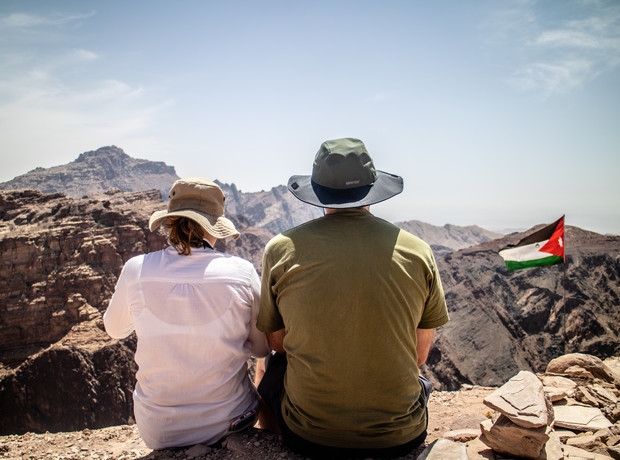 Two people sit on a rocky ledge, wearing hats and facing distant mountains; a Jordanian flag is visible in the swirling hot, arid landscape.