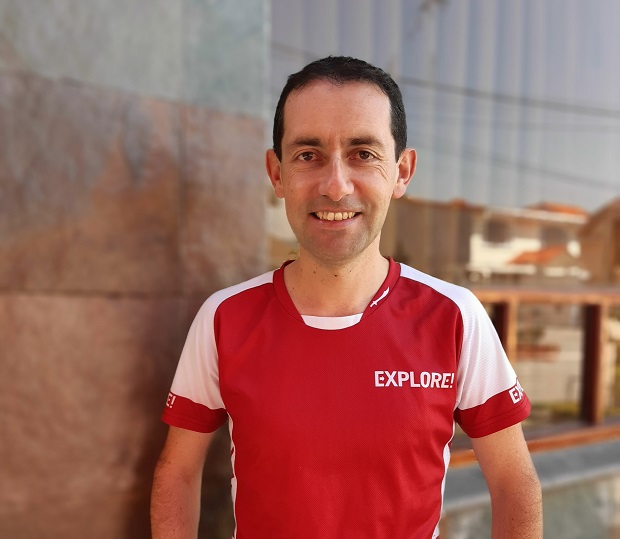 A person stands smiling, wearing a red and white shirt with the text "EXPLORE!" printed on it, near a stone and glass wall, in an urban setting.