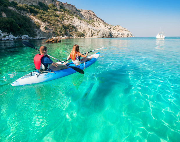 Two people paddle a kayak through clear, turquoise water, surrounded by rocky cliffs and distant cliffs. A white boat is visible on the horizon under a clear blue sky.