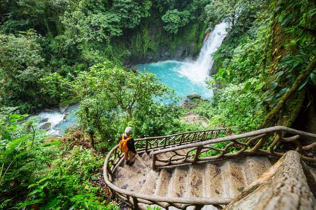 A person descends stone stairs surrounded by lush green forest, while a waterfall cascades into a vibrant turquoise pool below. The scene is serene and natural.