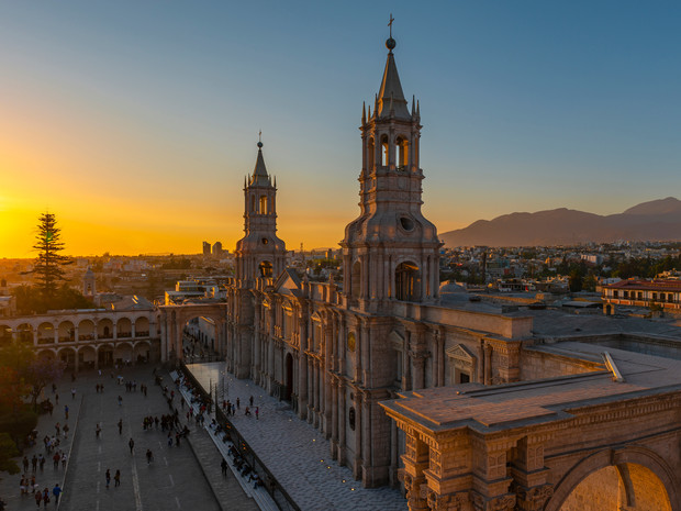 A cathedral with two prominent towers stands majestically at sunset, casting long shadows over a busy plaza in an urban landscape framed by distant mountains.