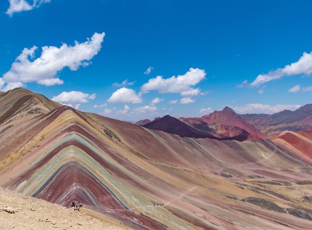 Mountain with colorful striations stands prominently; people hike along the ridge. The landscape extends under a bright blue sky with scattered clouds, creating a vibrant, natural setting.