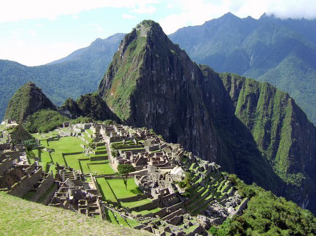 Ancient stone ruins rest amid green terraces, set against steep mountain peaks under a partly cloudy sky, suggesting a historical site nestled in a lush, mountainous area.