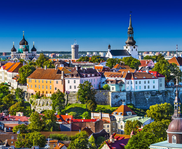 Historic buildings with colorful rooftops stand amidst lush green trees; church spires rise above, set against a deep blue sky in a charming urban landscape, suggesting a European cityscape.