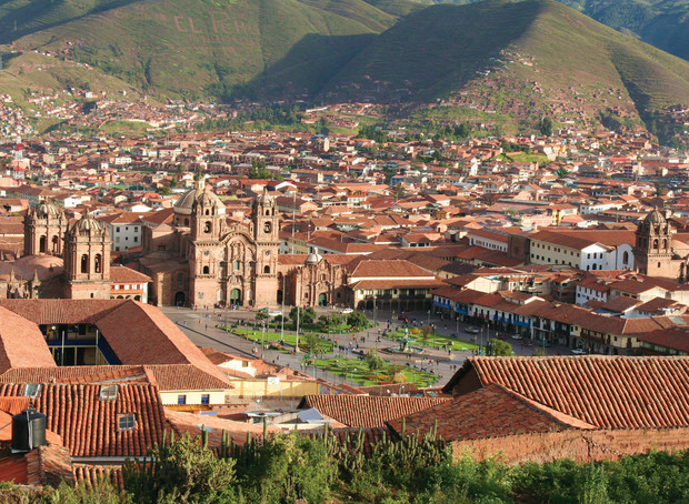 A historic cathedral stands prominently amidst a bustling, sunlit city square surrounded by red-roofed buildings, with green hills in the backdrop. Text "EL PERU" is visible on the hillside.