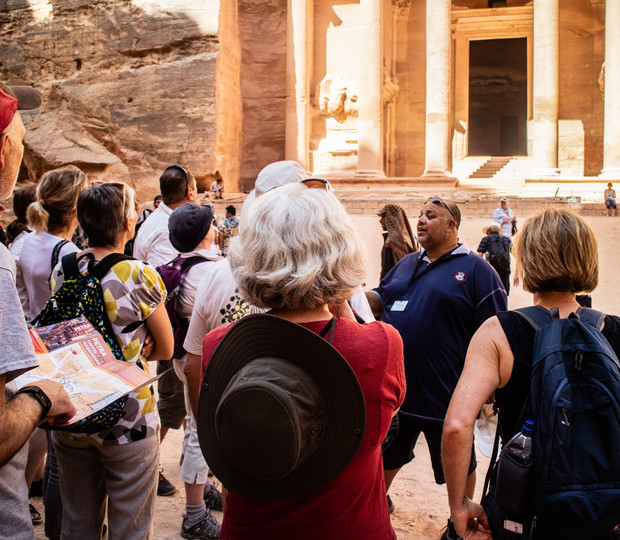 A tour guide talks to a group of tourists gathered in front of an ancient stone building with large columns, set within a sunlit desert landscape.