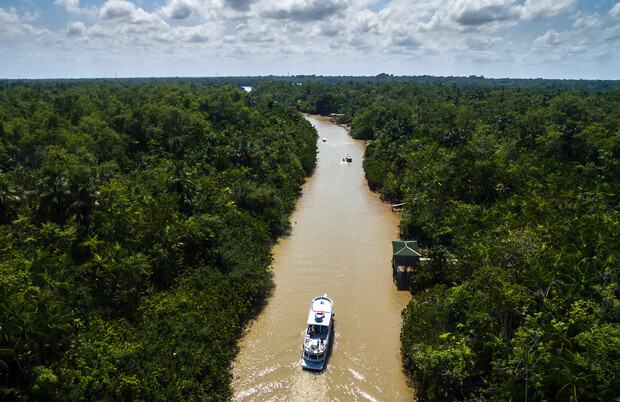 A boat travels along a narrow, winding river surrounded by dense green forest under a partly cloudy sky, with one small dock visible to the right.
