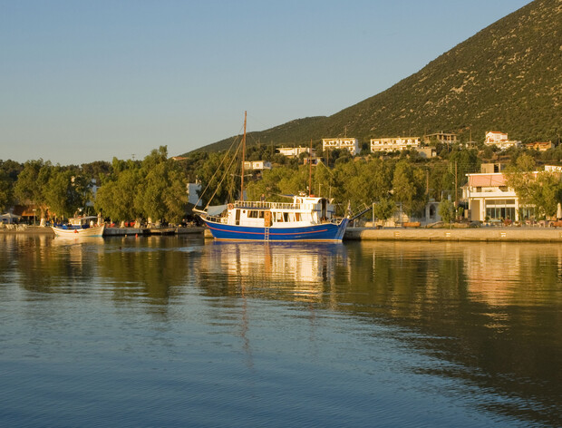 A blue and white boat docks at a tranquil waterfront, surrounded by trees and houses on a hillside under a clear sky.
