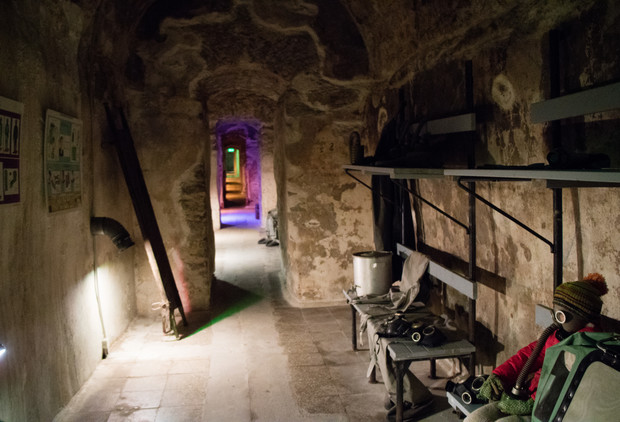 A narrow, dimly lit stone corridor features a bench with gas masks and a dressed mannequin. The walls are rough, and a green-lit open doorway leads to another area.