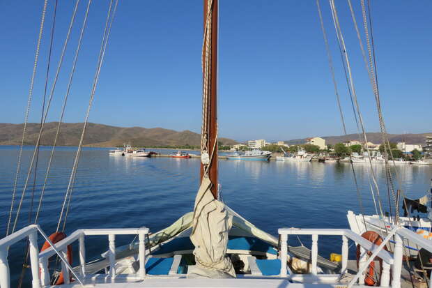 A boat's deck faces forward, with sails tied and mast upright, overlooking a tranquil harbor surrounded by distant hills and waterfront buildings under a clear blue sky.