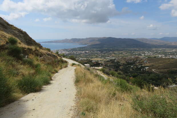 A winding dirt path leads downhill through grassy hills, overlooking a valley with scattered buildings and a distant blue coastline under a partly cloudy sky.
