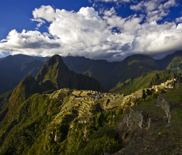 Ancient stone ruins sit atop a lush, green mountain ridge, set against dramatic cloud-covered peaks in the background, creating a serene and majestic landscape.