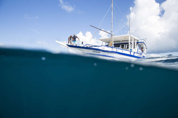 A boat floats on the ocean surface while people stand on deck. The image shows both above and below the waterline, revealing calm blue waters and a clear sky with clouds.