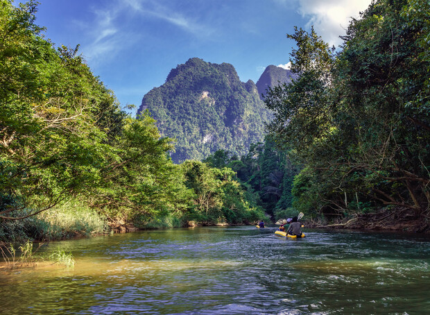 A kayaker navigates a serene river surrounded by lush, green trees, with towering mountains in the background under a partly cloudy sky.