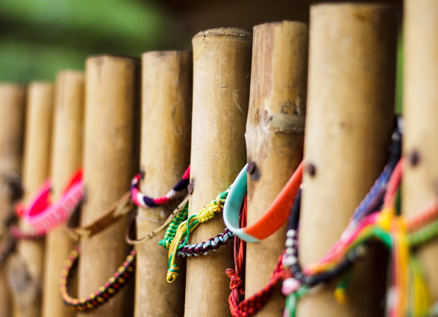 Colorful woven bracelets hang on vertical bamboo poles, creating a vibrant display. The scene appears outdoors, with a blurred green background suggesting a natural setting.