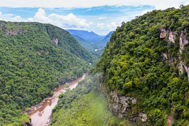 A river winds through a lush, green canyon with steep cliffs covered in dense forest, under a partly cloudy sky, creating a serene natural landscape.