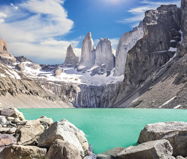 Jagged granite peaks rise dramatically behind a vivid turquoise lake, surrounded by rugged rocky terrain and patches of snow, under a bright blue sky with scattered clouds.