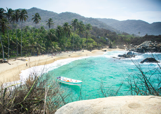 A small boat rests anchored near a sandy beach, bordered by palm trees and lush vegetation, with mountains in the background and several people walking along the shoreline.