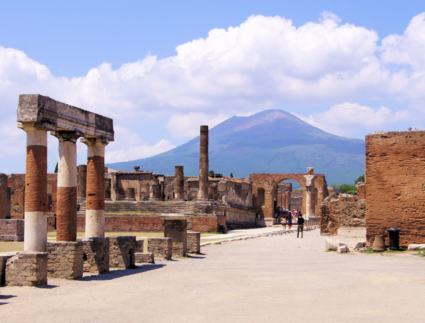 Ancient stone columns and ruins stand along a dusty pathway, with people walking in the distance. In the background, a mountain rises against a sky filled with scattered clouds.